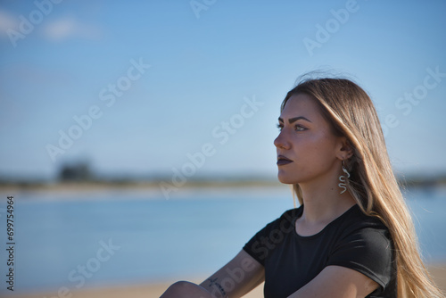Portrait of young, beautiful blonde woman, green eyes, with black top and tattoos, sitting, looking at infinity with the sea in the background. Concept of peace, tranquility, relaxation.