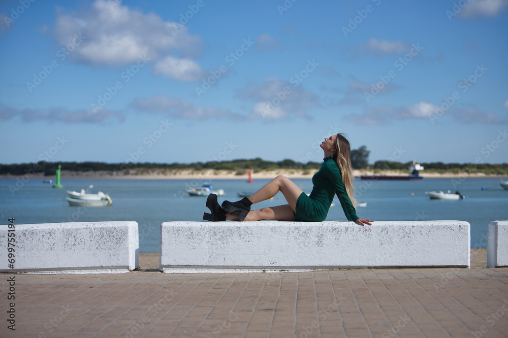 Young, beautiful, blonde woman, eyes closed and wearing a green dress, sitting on a stone bench, solitary, relaxed and calm, with the sea in the background. Concept of solitude, peace, tranquility.