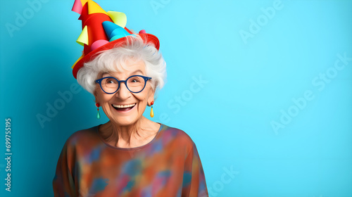Funny old senior woman or grandma wearing a birthday hat, smiling and looking to the side. Aged female with glasses, funny elderly retired pensioner with gray hair and wrinkles. Blue wall background