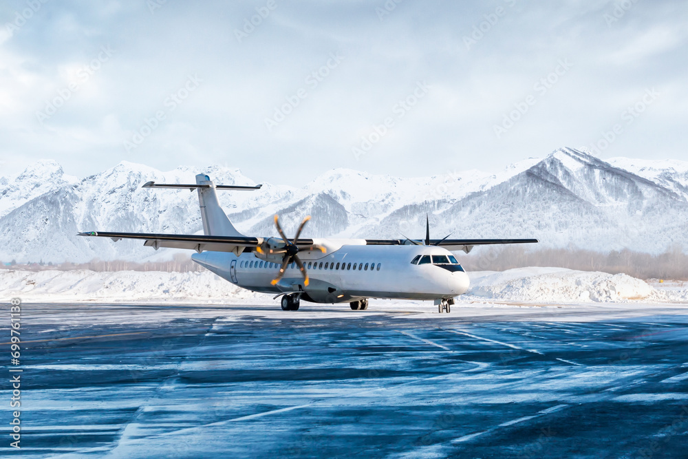 Naklejka premium White passenger turboprop airliner on the winter airport apron on the background of high picturesque mountains