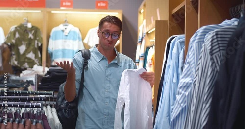 Man in men's clothing store closely examining shirt options Choice of shirt becomes challenging with high price tags. Surprise at cost during shirt selection moment of quality versus value. photo