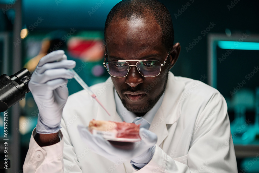 Black scientist dripping pink liquid on lab-grown meat sample sitting at his workplace in lab Black scientist dripping pink liquid on lab-grown meat sample sitting at his workplace in lab