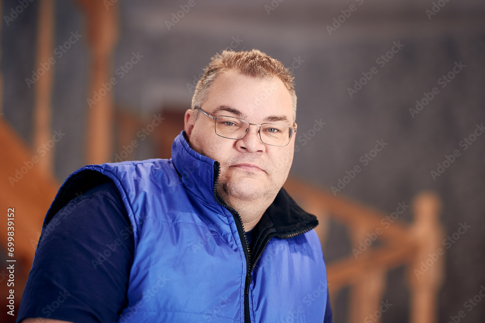 Portrait of sad, overweight man in his 40s wearing glasses and sleeveless jacket at home.