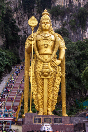 A majestic golden statue stands at the entrance to a cave with stairs and visitors in the background