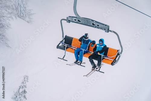 Skiers in ski suits and goggles ride on a chairlift above the snowy trees photo