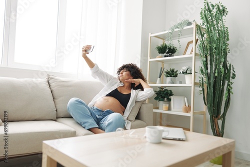 Pregnant woman blogger sits on the couch at home and takes pictures of herself on the phone, selfie and video call, consultation with the doctor online, pregnancy management