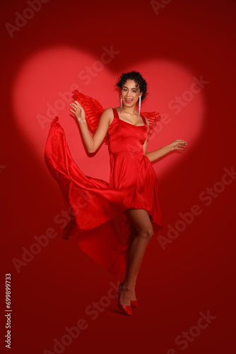 Young African-American woman dressed as Cupid on red background. Valentine's Day celebration