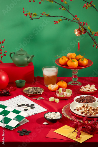 On a vibrant red and green backdrop, a go chess board, a glass of soft drink, plates of tangerines, sunflower seeds, colorful gummy candy, and a tea set create a diverse and enticing display. photo