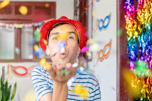 Brazilian man cheerfully prepares for Carnival at home, blowing confetti from his palm towards the camera. Captures the vibrant spirit and festive atmosphere