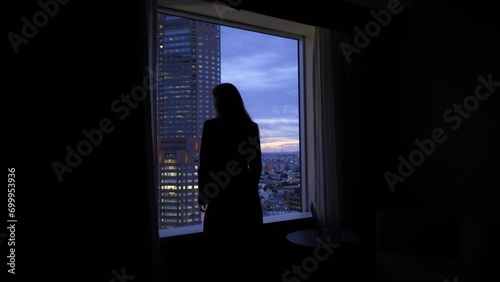 Woman come to window and look out to evening city, dark room, silhouetted figure against window, view from back and side. Lady wear informal suit. Blurred Tokyo cityscape and tall tower on back