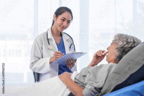 Elderly Asian woman lying in hospital bed looking unwell, with young Asian doctor take a history of illness Health check and encouragement.