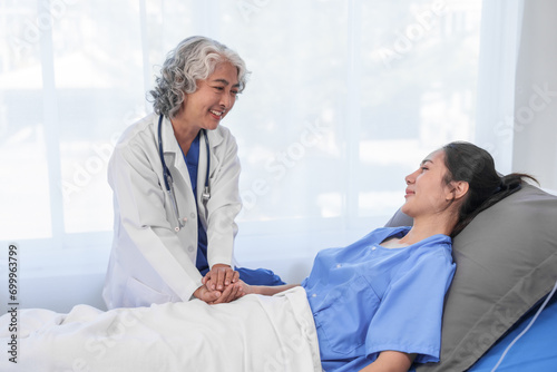 An elderly Asian doctor is smiling and holding the hand of a younger Asian patient who is lying in a hospital bed.