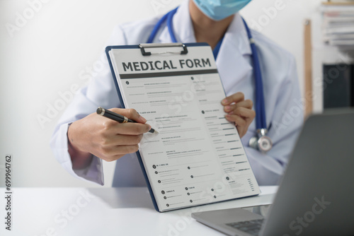 A doctor in a white coat and stethoscope is holding a medical form and a pen, with a laptop in the background.