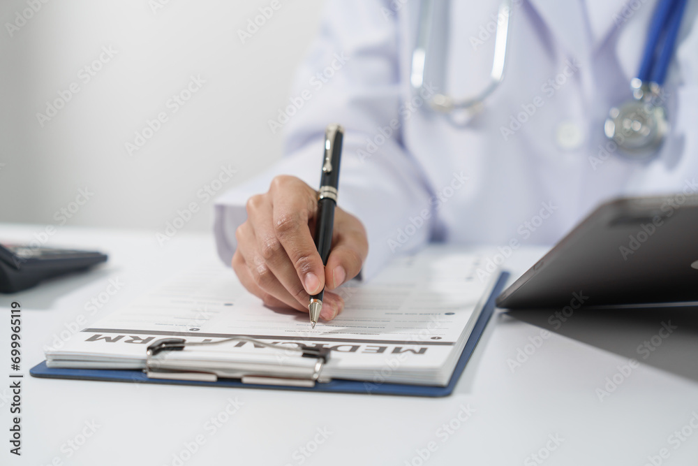 Fototapeta premium A doctor's hand is writing on a medical record form on a clipboard, with a stethoscope and tablet in the background.