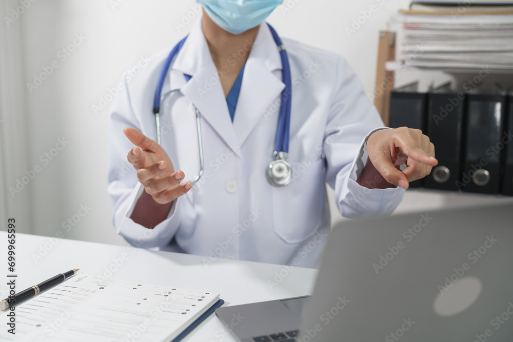 A doctor in a white coat with a stethoscope and face mask is gesturing and explaining something during an online consultation via laptop.