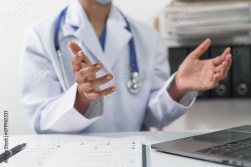 A doctor in a white coat with a stethoscope and face mask is gesturing and explaining something during an online consultation via laptop.