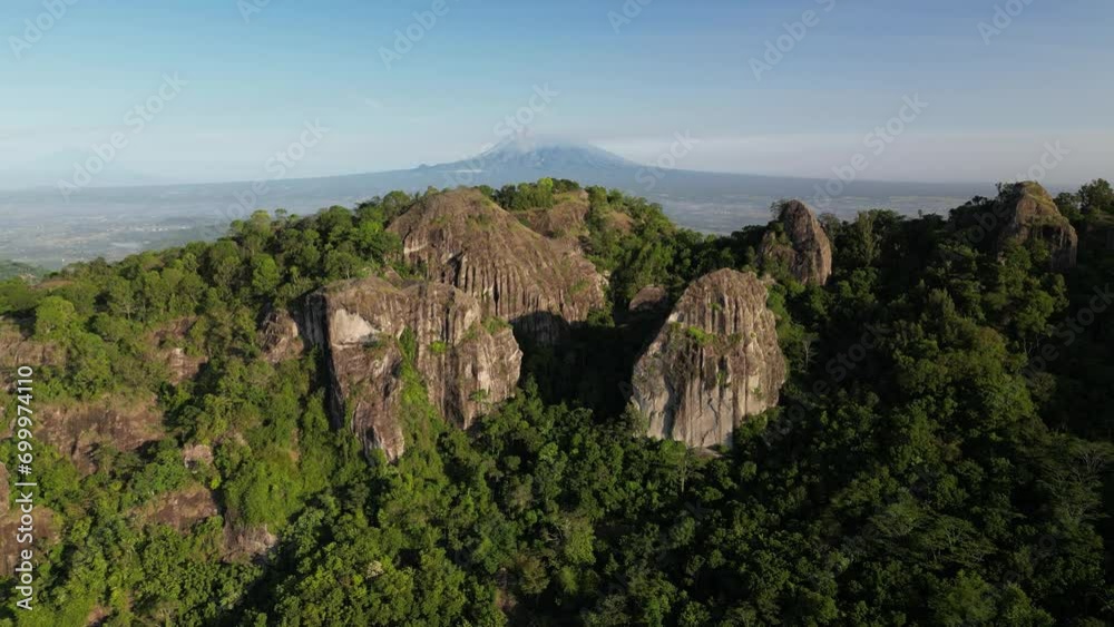 Mount Merapi seen afar from the peak of the Nglanggeran Ancient Volcano, Gunungkidul, Yogyakarta with clear blue sky background. The volcano is visible from behind the rocky mountains and forest. Mount Merapi seen afar from the peak of the Nglanggeran Ancient Volcano, Gunungkidul, Yogyakarta with clear blue sky background. The volcano is visible from behind the rocky mountains and forest.