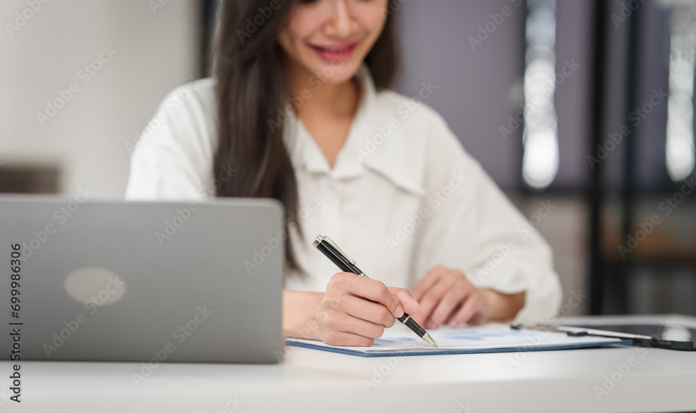 young Asian business woman with long hair writing on paper at a desk with a laptop open in front of her. young Asian business woman with long hair writing on paper at a desk with a laptop open in front of her.