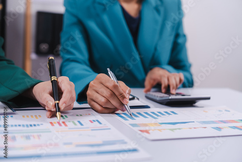 Two professionals are calculating finances, one using a calculator and the other taking notes, surrounded by colorful graphs and charts. photo