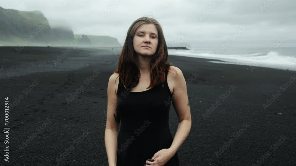Young beautiful woman in black dress at black sand beach Iceland, looking into camera model posing, slow motion, dramatic waves seascape	