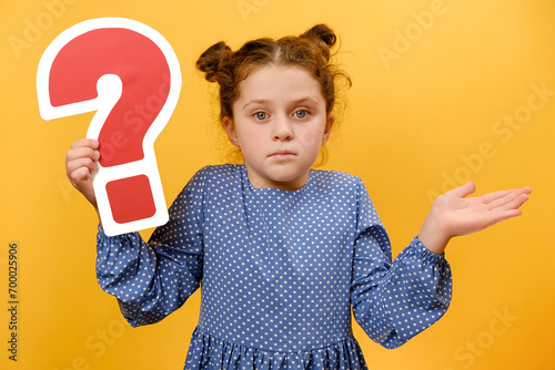 Portrait of pensive little girl kid 7-8 old years holding red question mark sign, confusion concept, posing isolated over plain yellow color background wall in studio. Childhood lifestyle concept