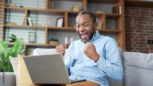 Happy excited mature african american man received great news on laptop sitting on sofa in living room at home. Smiling senior bearded male reads a pleasant message on the computer, celebrates success photo