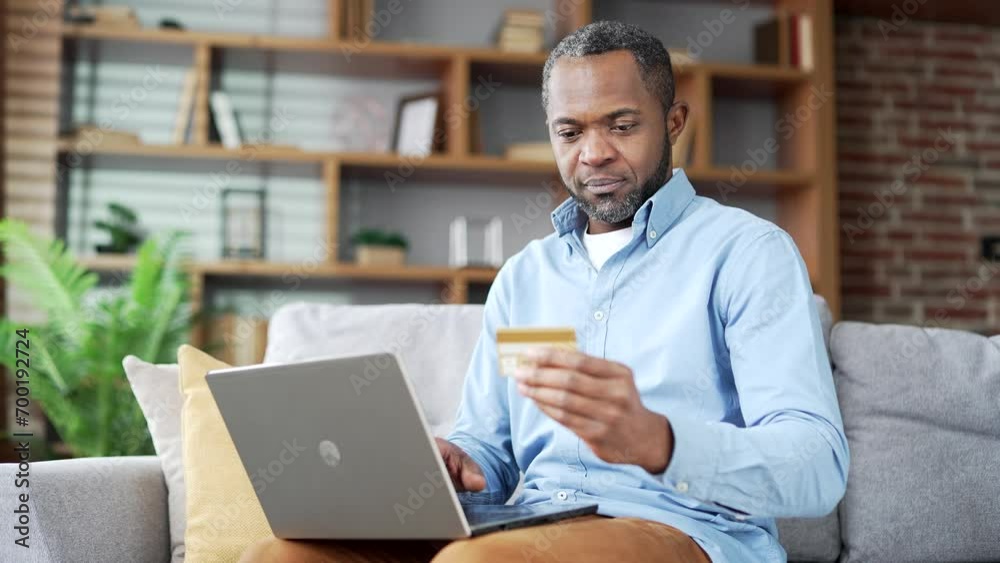 Happy mature african american man doing online shopping typing credit card number on laptop sitting on sofa at home. Smiling senior bearded male makes purchase, satisfied with promotions and discounts
