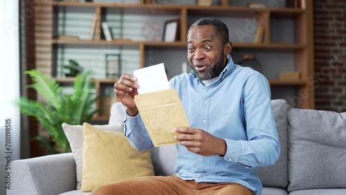 Happy excited mature african american man reading letter with great news sitting on sofa in living room at home. Joyful senior bearded male is satisfied with pleasant notification, celebrates success photo