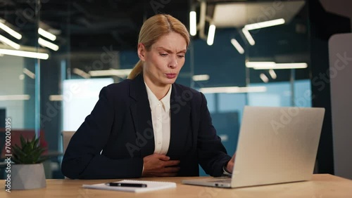 Sick ill woman suffering from stomach ache sitting at workplace, holding belly, feeling abdominal or menstrual pain indoor. Office worker having pain, gastritis, diarrhea, painful periods concept.