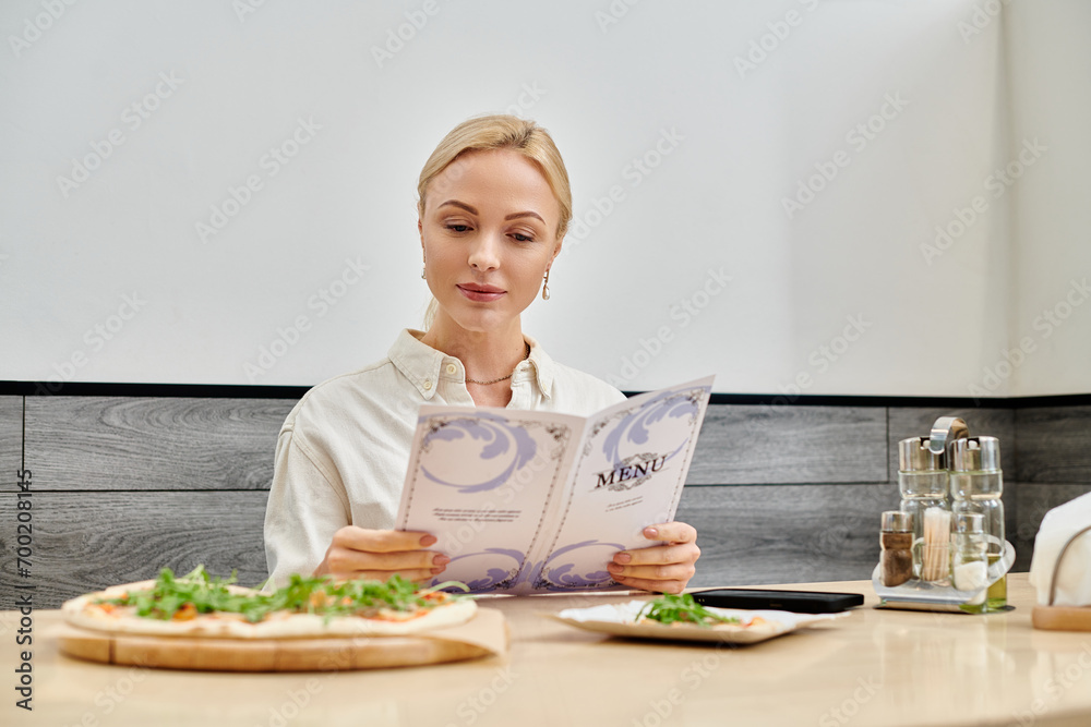 pretty blonde woman looking in menu card near delicious pizza while sitting in modern cafe
