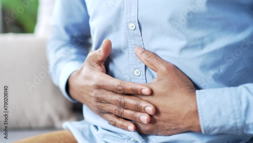 Close up. Male hands holding massaging stomach. Mature african american man in blue shirt feels stomach pain sitting on sofa in living room at home. Sick man suffers from bloating, gastritis or spasms photo