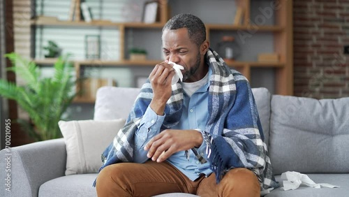 Sick mature african american male suffers from a runny nose sitting on sofa in the living room at home. Senior bearded black man wipes his nose with a handkerchief. He has a cold, fever, virus or flu photo