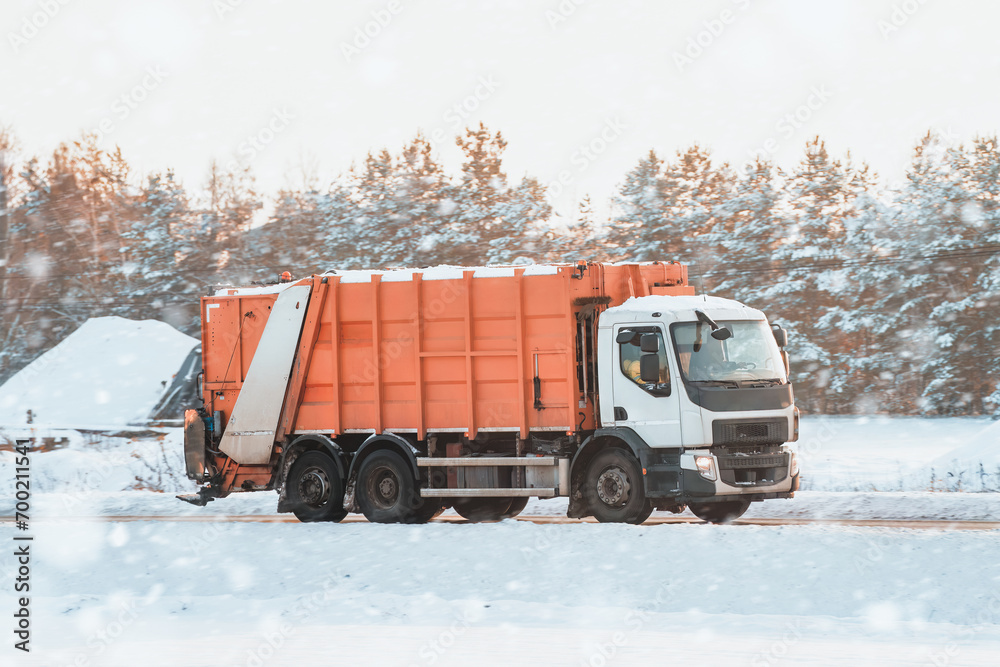 An orange sanitation truck moves on a snow-covered winter road. Garbage truck outdoor. Recycling truck rides on the road. An orange sanitation truck moves on a snow-covered winter road. Garbage truck outdoor. Recycling truck rides on the road.
