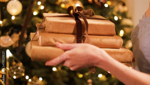 Close up of women hands giving and receiving Christmas gifts on background of decorated fir tree. Lady wearing stylish evening dress presenting pile of books tied with ribbon to her best friend. photo