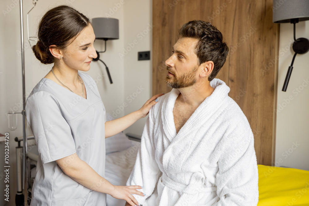 Fototapeta premium Young nurse takes care of adult man sitting on bed of comfortable medical ward. Medical support and assistance during the rehabilitation in clinic