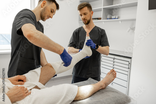 Two medical workers make a bandage on patient's leg in the trauma department. Concept of traumatology, orthopedics and rehabilitation after injuries