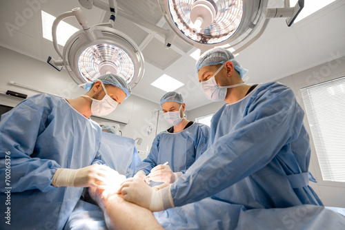 Three confident surgeons performing surgical operation on a patient's knee in operating room. Concept of real surgery and invasive treatments photo