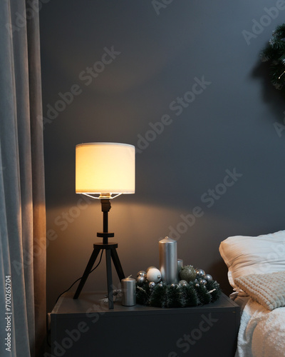 There is a nightstand in the room next to the bed, a nightlight on the nightstand. Gray wall, lamp and bed, hotel rest