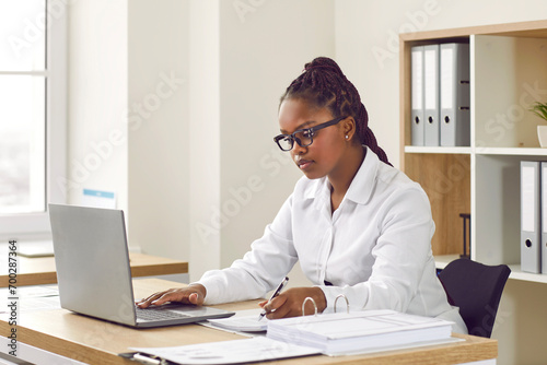 Focused African woman working on laptop at her desk in modern office. Confident attractive young woman in white blouse writing down notes on paper while looking at screen of computer photo