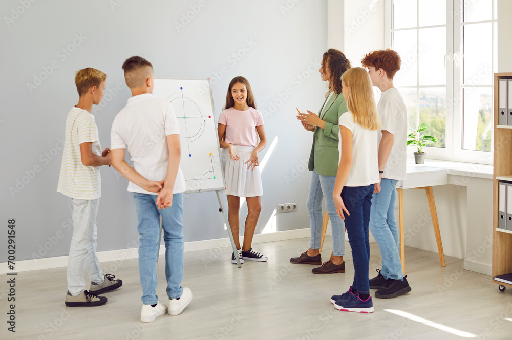 Naklejka premium Group of school children standing in a circle discussing study project with female friendly teacher. Student girl presenting project homework during class lesson in the classroom. Education concept.