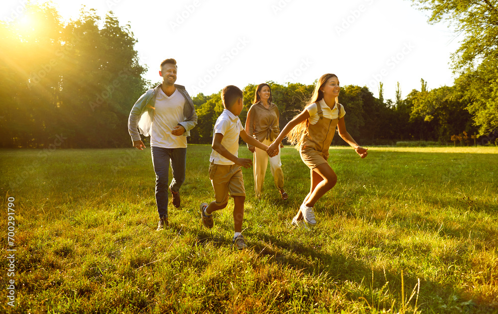 Cheerful young family with two children having fun and playing in summer park. Brother and sister holding hands run ahead of mom and dad on green grass in sunlight. Family leisure concept.