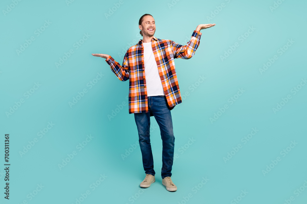 Full size photo of optimistic man dressed checkered shirt look at arm recommend product empty space isolated on teal color background Full size photo of optimistic man dressed checkered shirt look at arm recommend product empty space isolated on teal color background