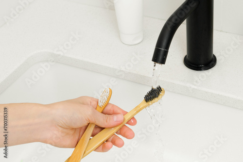 A woman's hand washes wooden bamboo toothbrushes from toothpaste after brushing teeth under a stream of water over a sink. Concept of oral hygiene and natural cosmetic products, sustainable lifestyle