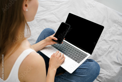 Young woman holding phone and laptop with blank black screen while sitting on sofa. Background mockup for your advertising content. The concept of remote work and watching online courses and movies.