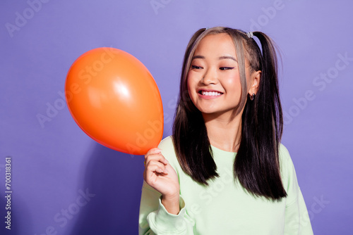 Portrait of youth girl korean person wearing trendy green jumper playing with orange helium air balloon isolated on violet color background © deagreez