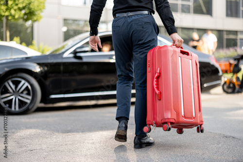 Man carries red suitcase to a car, cropped view from below. Concept of chauffeur service and business trips