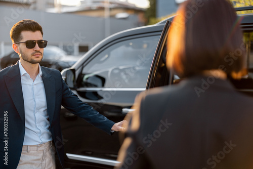 Elegant business woman walks to the luxury car, man with a suitcase opens vehicle door, letting lady in. Concept of business trip and travel