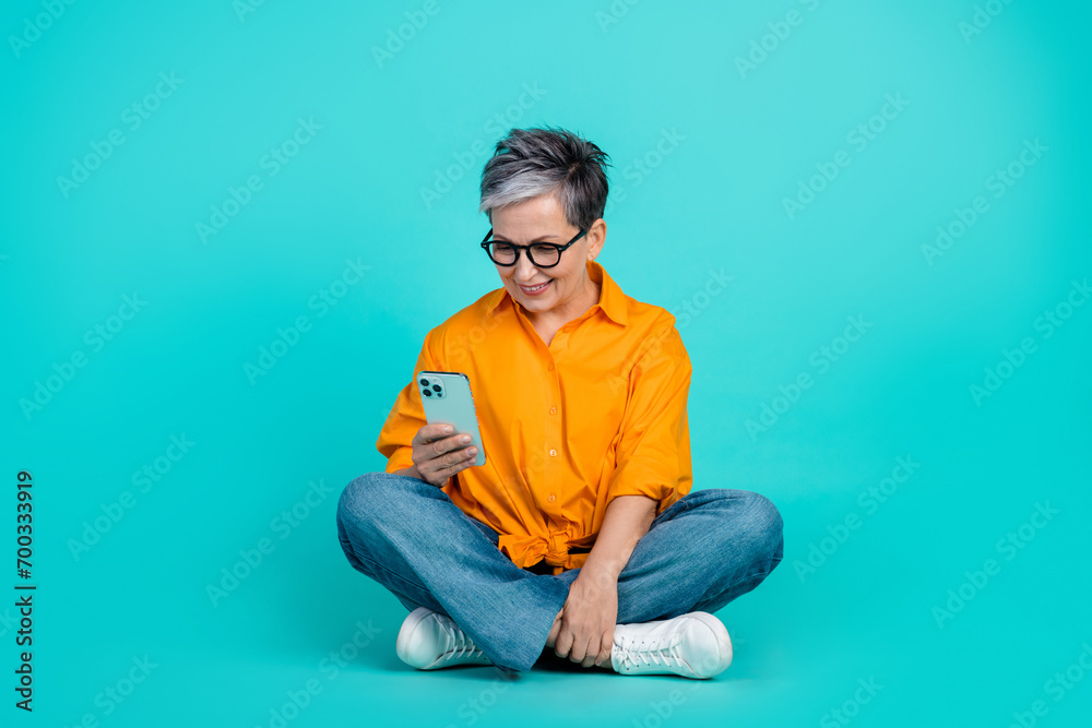 Full body size photo of focused working business woman pensioner browsing smartphone sit floor isolated on aquamarine color background Full body size photo of focused working business woman pensioner browsing smartphone sit floor isolated on aquamarine color background