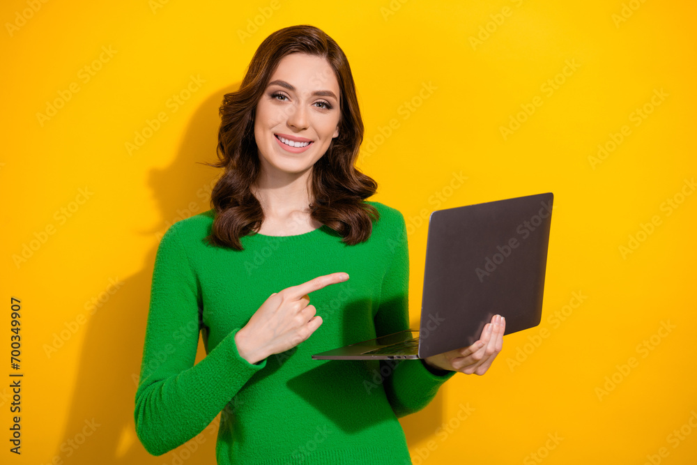 Portrait of pretty positive girl beaming smile indicate finger arm hold netbook isolated on yellow color background Portrait of pretty positive girl beaming smile indicate finger arm hold netbook isolated on yellow color background