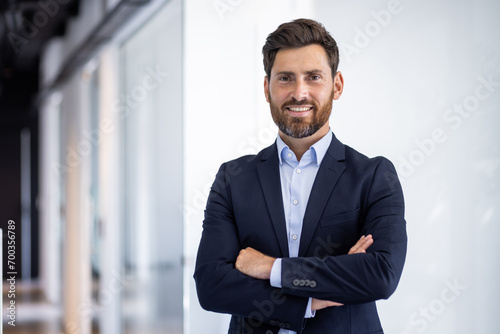 Portrait of a smiling young successful male businessman standing in the office in a suit and looking at the camera with his arms crossed on his chest photo